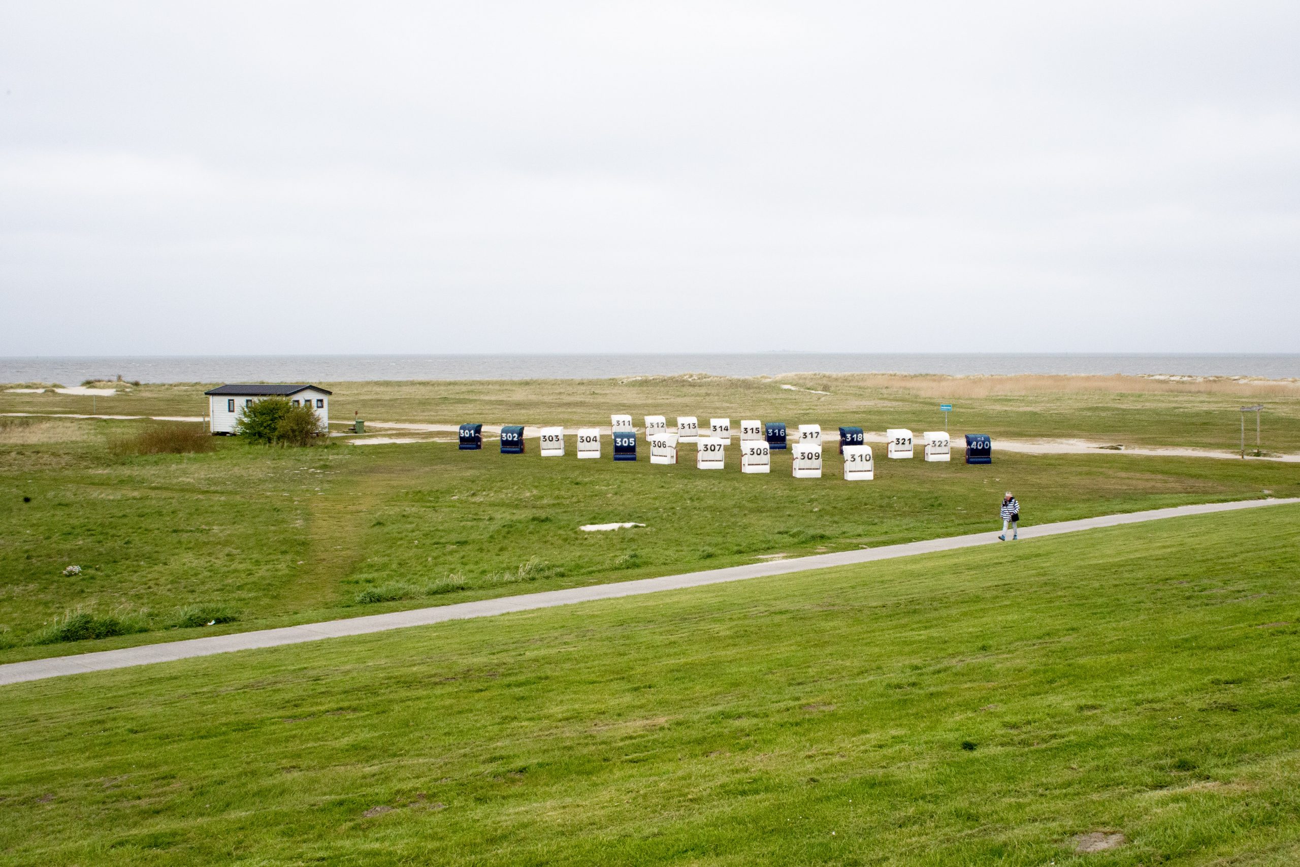 Eine klassische Nordseestrand-Szenerie mit einer langen Reihe nummerierter Strandkörbe in Weiß, Blau und anderen Farben (Nummern 310-400+). Die robusten Flechtwerk-Körbe stehen auf grüner Strandwiese. Ein gepflasterter Weg führt parallel vorbei, auf dem Fußgänger spazieren. Ein weißes charakteristisches Strandhaus mit grünem Dach sitzt links in der Szene. Der Horizont zeigt das ruhige Meer unter grauem Himmel. Die Szenerie vermittelt klassisches deutsches Nordseebad-Flair mit Entspannung und maritimem Urlaubscharme.