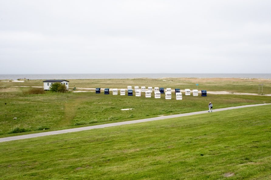 Eine klassische Nordseestrand-Szenerie mit einer langen Reihe nummerierter Strandkörbe in Weiß, Blau und anderen Farben (Nummern 310-400+). Die robusten Flechtwerk-Körbe stehen auf grüner Strandwiese. Ein gepflasterter Weg führt parallel vorbei, auf dem Fußgänger spazieren. Ein weißes charakteristisches Strandhaus mit grünem Dach sitzt links in der Szene. Der Horizont zeigt das ruhige Meer unter grauem Himmel. Die Szenerie vermittelt klassisches deutsches Nordseebad-Flair mit Entspannung und maritimem Urlaubscharme.