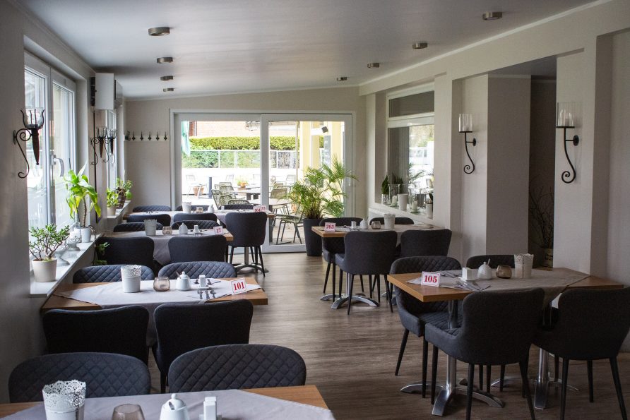 Helles, modernes Restaurantinterieur mit mehreren Tischen, die mit weißen Tischdecken gedeckt sind, schwarzen Stühlen und nummerierten Tischschildern. Große Fenster lassen Tageslicht herein; grüne Pflanzen schmücken den Raum. Eine Glastür im hinteren Bereich führt zu einer Sitzgelegenheit im Freien.