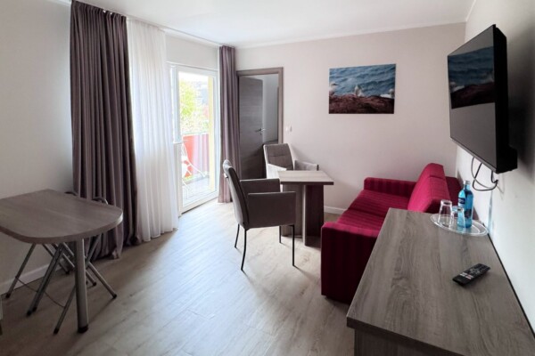 Eine elegante Suite mit Schlaf- und Wohnbereich. Das Doppelbett mit grauem Kissen befindet sich links neben großen Fenstern mit Balkonzugang. Der Wohnbereich rechts zeigt einen braunen Sessel, ein rotes Sofa und einen hölzernen Schreibtisch mit schwarzem Stuhl. Ein großer schwarzer Flachbild-TV hängt an der beige-rosa Wand. Ein Kunstwerk mit Meereslandschaft verstärkt die maritime Ästhetik. Der hellbraune Holzfußboden, doppelte Vorhänge und durchdachte Möblierung schaffen eine luxuriöse, funktionale Wohnatmosphäre für längere Aufenthalte. Eine elegante Suite mit Schlaf- und Wohnbereich. Das Doppelbett mit grauem Kissen befindet sich links neben großen Fenstern mit Balkonzugang. Der Wohnbereich rechts zeigt einen braunen Sessel, ein rotes Sofa und einen hölzernen Schreibtisch mit schwarzem Stuhl. Ein großer schwarzer Flachbild-TV hängt an der beige-rosa Wand. Ein Kunstwerk mit Meereslandschaft verstärkt die maritime Ästhetik. Der hellbraune Holzfußboden, doppelte Vorhänge und durchdachte Möblierung schaffen eine luxuriöse, funktionale Wohnatmosphäre für längere Aufenthalte.
