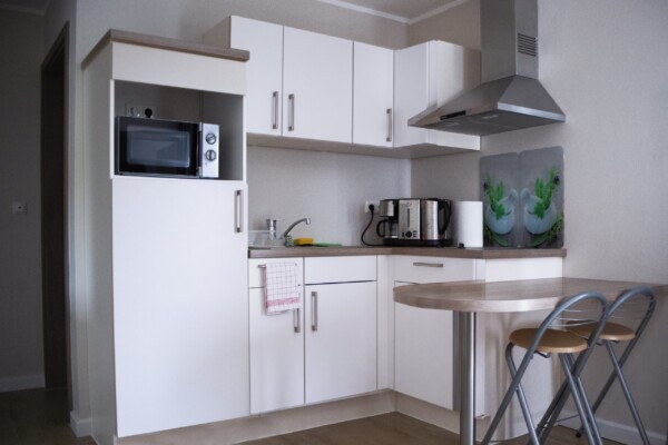 Eine moderne L-förmige Kitchenette mit weißen Hochglanz-Schränken und hölzernem Arbeitsplan in warmer Brauntönung. In einer dunklen Nische ist eine schwarze Mikrowelle integriert. Ein dunkles Kochfeld mit metallener Dunstabzugshaube komplettiert die Kochzone. Ein schwarzer Wasserkocher und eine Kaffemaschine stehen griffbereit auf dem Arbeitsplan. Grüne Topfpflanzen geben einen frischen Akzent. Ein massiver hölzerner Tisch mit zwei braunen Stühlen bildet den Essbereich. Die helle, funktionale Ausstattung schafft eine moderne, praktische Atmosphäre für Selbstversorger. Eine moderne L-förmige Kitchenette mit weißen Hochglanz-Schränken und hölzernem Arbeitsplan in warmer Brauntönung. In einer dunklen Nische ist eine schwarze Mikrowelle integriert. Ein dunkles Kochfeld mit metallener Dunstabzugshaube komplettiert die Kochzone. Ein schwarzer Wasserkocher und eine Kaffemaschine stehen griffbereit auf dem Arbeitsplan. Grüne Topfpflanzen geben einen frischen Akzent. Ein massiver hölzerner Tisch mit zwei braunen Stühlen bildet den Essbereich. Die helle, funktionale Ausstattung schafft eine moderne, praktische Atmosphäre für Selbstversorger.