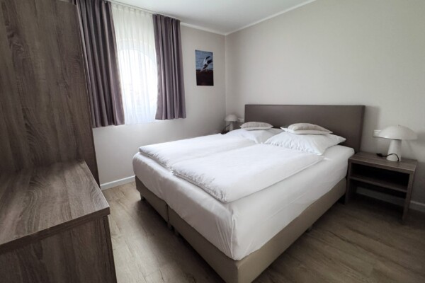 Ein schlichtes, modern ausgestattetes Schlafzimmer mit grauem Doppelbett und weißer Bettwäsche. Der hellbraune Holzfußboden erstreckt sich durchgehend. Große Fenster mit graubraunen und weißen Doppelvorhängen ermöglichen reichlich Tageslicht. Ein dunkler Nachttisch mit eleganter weißer Tischleuchte flankiert das Bett. Ein kleines Berglandschaftsbild hängt dezent an der Wand. Die minimalistische Einrichtung und neutrale Farbpalette schaffen eine ruhige, entspannende Atmosphäre ohne Ablenkung. Ein schlichtes, modern ausgestattetes Schlafzimmer mit grauem Doppelbett und weißer Bettwäsche. Der hellbraune Holzfußboden erstreckt sich durchgehend. Große Fenster mit graubraunen und weißen Doppelvorhängen ermöglichen reichlich Tageslicht. Ein dunkler Nachttisch mit eleganter weißer Tischleuchte flankiert das Bett. Ein kleines Berglandschaftsbild hängt dezent an der Wand. Die minimalistische Einrichtung und neutrale Farbpalette schaffen eine ruhige, entspannende Atmosphäre ohne Ablenkung.