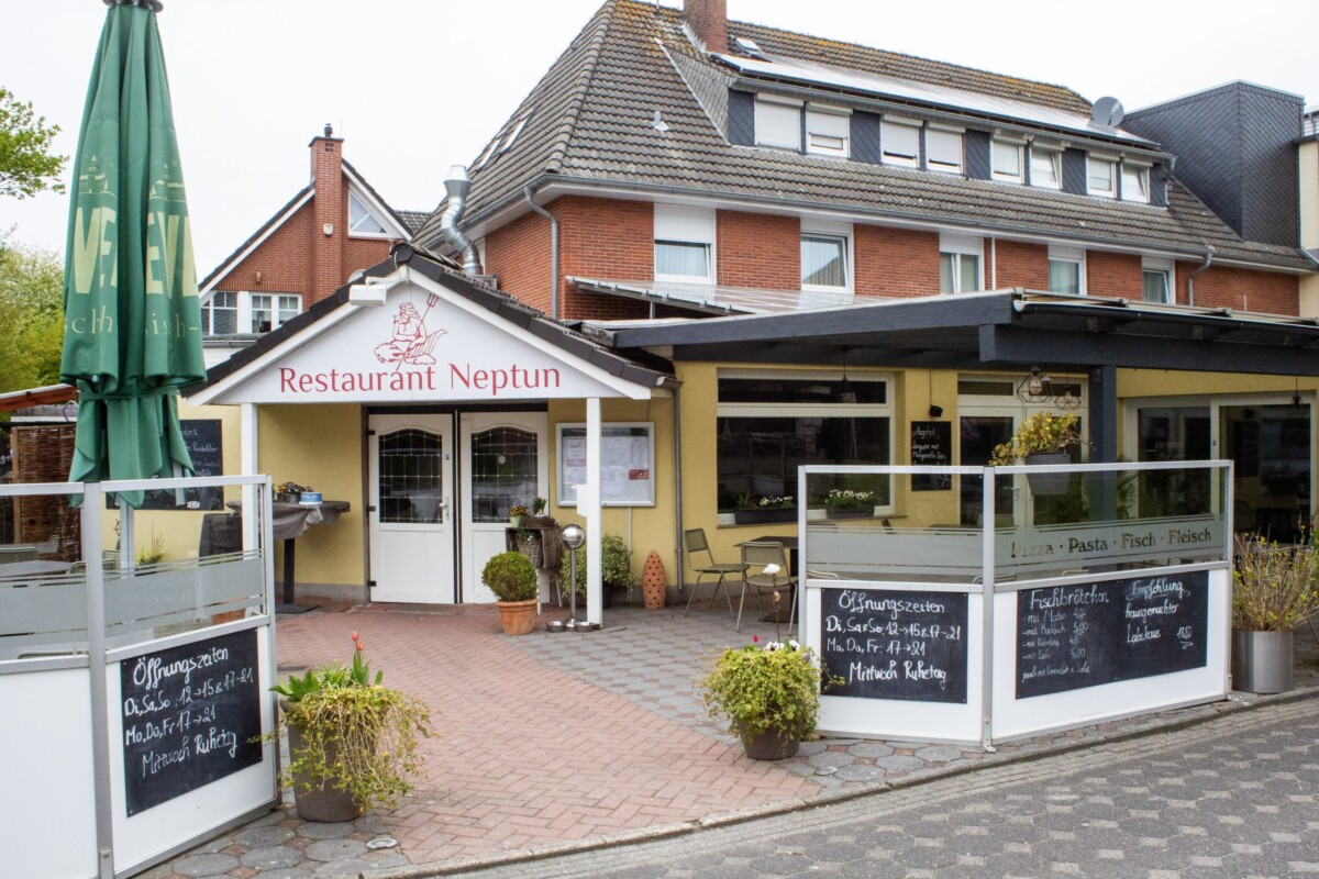 Das Restaurant Neptun präsentiert sich mit einer charming restaurierten Gelbfassade und traditionellem Ziegeldach. Das rustikale Backsteingebäude dahinter bildet einen authentischen Rahmen. Die Terrasse ist mit weißen Gardinen, grünen Sonnenschirmen und Topfpflanzen liebevoll gestaltet. Mehrere Schiefertafeln-Menüboards mit handschriftlichen Einträgen zeigen Pasta-, Fisch- und Fleischspezialitäten sowie Öffnungszeiten. Tische und Stühle laden zum Verweilen ein. Die gemütliche, traditionelle Gasthof-Atmosphäre mit grünem Bewuchs wirkt einladend und authentisch-regional.