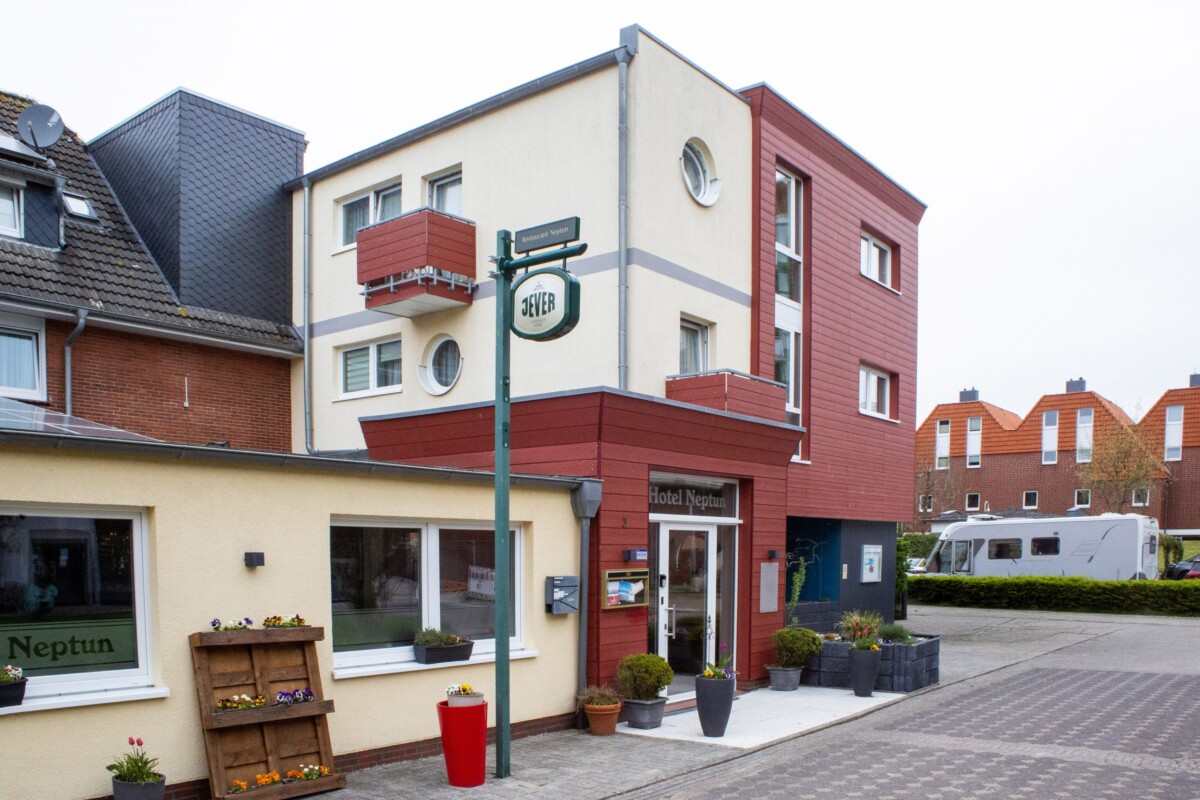 Das Hotel Neptun präsentiert sich mit moderner, gemischter Architektur: Die oberen Stockwerke zeigen rote Holzverkleidung mit regulären Fenstern, während die unteren Bereiche eine cremefarbene Fassade mit weißen Fensterrahmen aufweisen. Ein grünes metallisches Schild mit der Hotelbezeichnung prangt seitlich. Die Eingangstür ist in warmen Rotbraun-Tönen gehalten, flankiert von gepflegten Blumenkästen und Topfpflanzen. Eine holzerne Blumenpyramide mit bunten Blüten begrüßt Gäste. Die urbane Lage, die durchdachte Begrünung und die freundliche Fassadengestaltung schaffen ein einladendes, modernes Hotelambiente.