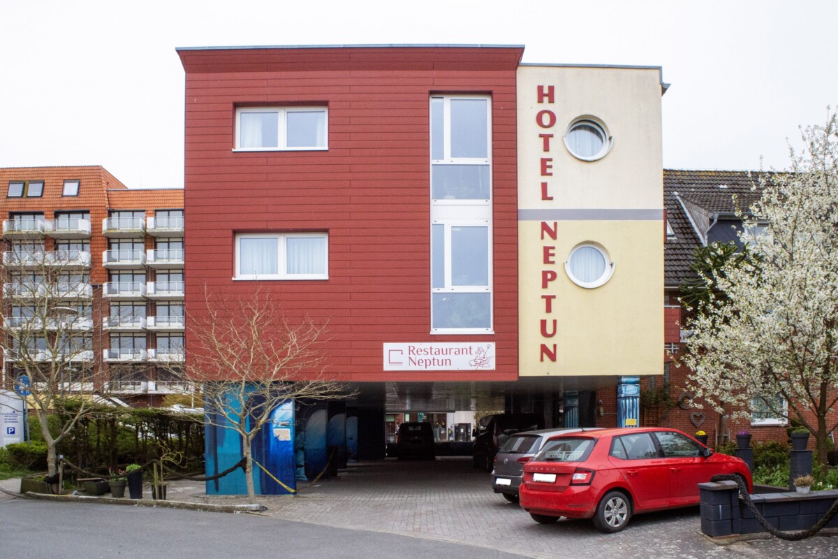 Das Hotel Neptun dominiert die Straßenszene mit einer imposanten roten Holzfassade und großflächigen Fenstern. Ein cremefarbener Gebäudeteil trägt die vertikale Beschriftung 'HOTEL NEPTUN' in roter Schrift. Das Restaurant Neptun befindet sich im Erdgeschoss mit dunklem Eingangsbereich. Mehrere Wohngebäude mit rotem Klinker flankieren das Hotel links. Ein rotes Auto parkt vor dem Eingang auf dem Kopfsteinpflaster. Die urbane Lage und moderne Architektur schaffen ein einladendes Hotel-Erscheinungsbild.