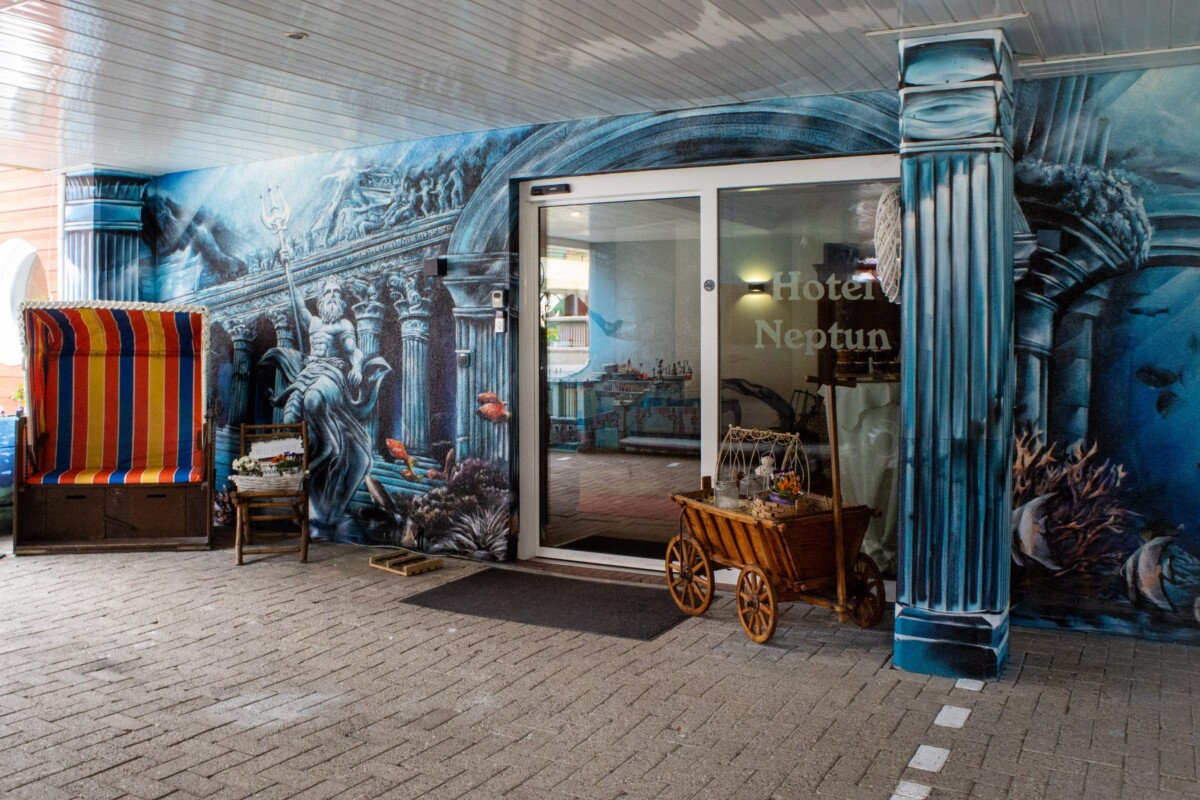 Eine großformatige künstlerische Wandmalerei ziert den Eingangsbereich des Hotels mit klassischen mythologischen Neptun-Motiven. Die blautonige Wandgestaltung zeigt detaillierte Skulpturfiguren und architektonische Elemente. Eine rot-gelb gestreifte Vintage-Sonnenliege und ein charmanter Holzwagen ergänzen die maritime Thematik. Eine gläserne Eingangstür mit 'Hotel Neptun' Beschriftung führt ins Innere. Der künstlerische Wandaufbau schafft eine einprägsame, kulturell bereichernde Willkommens-Atmosphäre für Besucher.