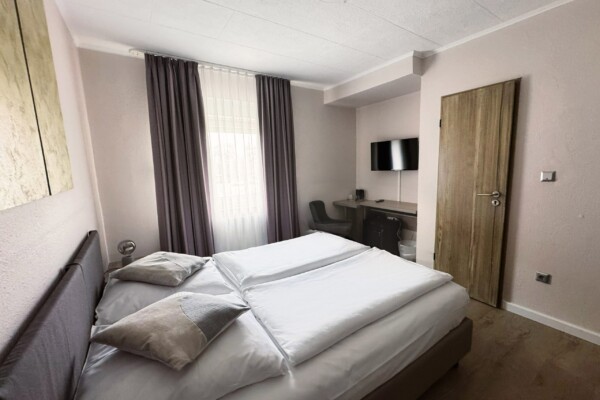 Ein kleines, modernes Hotelzimmer mit einem ordentlich gemachten Doppelbett, zwei Kissen, hellen Wänden, einem Fenster mit transparenten und dunklen Vorhängen, einer Holztür, einem Schreibtisch mit Stuhl, einem an der Wand montierten Flachbildfernseher und einem Bodenbelag in Holzoptik. Ein kleines, modernes Hotelzimmer mit einem ordentlich gemachten Doppelbett, zwei Kissen, hellen Wänden, einem Fenster mit transparenten und dunklen Vorhängen, einer Holztür, einem Schreibtisch mit Stuhl, einem an der Wand montierten Flachbildfernseher und einem Bodenbelag in Holzoptik.