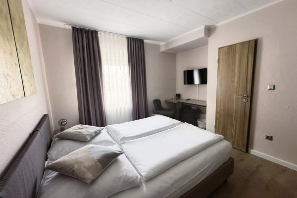 Ein kleines, modernes Hotelzimmer mit einem ordentlich gemachten Doppelbett, zwei Kissen, hellen Wänden, einem Fenster mit transparenten und dunklen Vorhängen, einer Holztür, einem Schreibtisch mit Stuhl, einem an der Wand montierten Flachbildfernseher und einem Bodenbelag in Holzoptik. Ein kleines, modernes Hotelzimmer mit einem ordentlich gemachten Doppelbett, zwei Kissen, hellen Wänden, einem Fenster mit transparenten und dunklen Vorhängen, einer Holztür, einem Schreibtisch mit Stuhl, einem an der Wand montierten Flachbildfernseher und einem Bodenbelag in Holzoptik.