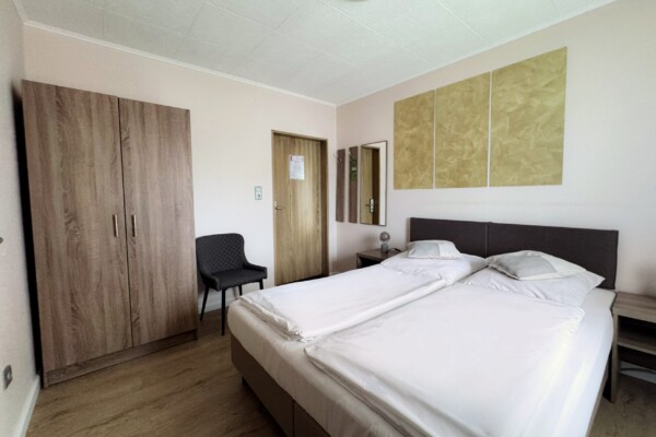 Ein aufgeräumtes Hotelzimmer mit einem Doppelbett, zwei Kissen und weißer Bettwäsche. Links steht ein Kleiderschrank aus Holz, daneben ein Stuhl, und hinten befindet sich eine Tür. Über dem Bett hängen drei goldene Tafeln, und zwei Nachttische mit Lampen flankieren das Bett. Ein aufgeräumtes Hotelzimmer mit einem Doppelbett, zwei Kissen und weißer Bettwäsche. Links steht ein Kleiderschrank aus Holz, daneben ein Stuhl, und hinten befindet sich eine Tür. Über dem Bett hängen drei goldene Tafeln, und zwei Nachttische mit Lampen flankieren das Bett.