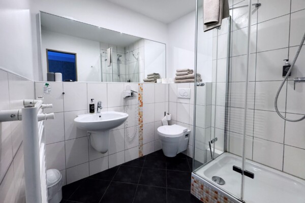 Modernes Badezimmer mit weißen Fliesenwänden und schwarzem Fliesenboden. Ausgestattet mit einem wandmontierten Waschbecken, einer Dusche mit Glastür, einer Toilette, einem großen Spiegel, gestapelten Handtüchern, einem Handtuchheizkörper, einem Toilettenpapierhalter, einem Seifenspender und einer Deckenbeleuchtung. Modernes Badezimmer mit weißen Fliesenwänden und schwarzem Fliesenboden. Ausgestattet mit einem wandmontierten Waschbecken, einer Dusche mit Glastür, einer Toilette, einem großen Spiegel, gestapelten Handtüchern, einem Handtuchheizkörper, einem Toilettenpapierhalter, einem Seifenspender und einer Deckenbeleuchtung.