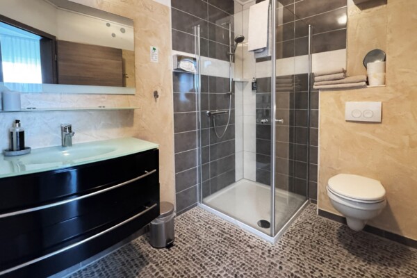 Modernes Badezimmer mit einer Duschkabine aus Glas, einem wandhängenden WC, einem schwebenden schwarzen Waschtisch mit Glasplatte und Regalen für Handtücher. Der Boden weist eine kieselsteinartige Struktur auf, und die Wände sind mit beigen und dunkelgrauen Fliesen verkleidet. Modernes Badezimmer mit einer Duschkabine aus Glas, einem wandhängenden WC, einem schwebenden schwarzen Waschtisch mit Glasplatte und Regalen für Handtücher. Der Boden weist eine kieselsteinartige Struktur auf, und die Wände sind mit beigen und dunkelgrauen Fliesen verkleidet.