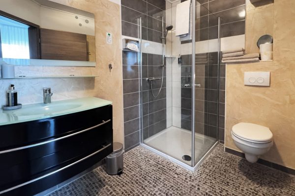 Modernes Badezimmer mit einer Duschkabine aus Glas, einem wandhängenden WC, einem schwebenden schwarzen Waschtisch mit Glasplatte und Regalen für Handtücher. Der Boden weist eine kieselsteinartige Struktur auf, und die Wände sind mit beigen und dunkelgrauen Fliesen verkleidet. Modernes Badezimmer mit einer Duschkabine aus Glas, einem wandhängenden WC, einem schwebenden schwarzen Waschtisch mit Glasplatte und Regalen für Handtücher. Der Boden weist eine kieselsteinartige Struktur auf, und die Wände sind mit beigen und dunkelgrauen Fliesen verkleidet.