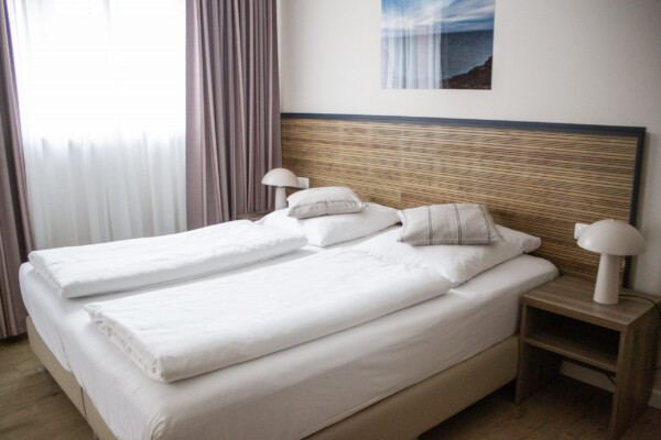 Ein modernes Hotelzimmer mit einem breiten Doppelbett, das weiße Bettwäsche und mehrere graue Kissen aufweist. Das Kopfbett ist aus hellem Holz mit horizontalen Rillen gefertigt. Neben dem Bett stehen hölzerne Nachttische mit weißen Pilzlampen. Große Fenster mit graubraunen Vorhängen und einer weißen Innenschicht ermöglichen reichlich Tageslicht. An der Wand hängt eine Berglandschaftsfotografie. Ein weißer und dunkelblauer Wandstreifen strukturiert den Raum. Die Atmosphäre wirkt modern, minimalistisch und einladend erholsam. Ein modernes Hotelzimmer mit einem breiten Doppelbett, das weiße Bettwäsche und mehrere graue Kissen aufweist. Das Kopfbett ist aus hellem Holz mit horizontalen Rillen gefertigt. Neben dem Bett stehen hölzerne Nachttische mit weißen Pilzlampen. Große Fenster mit graubraunen Vorhängen und einer weißen Innenschicht ermöglichen reichlich Tageslicht. An der Wand hängt eine Berglandschaftsfotografie. Ein weißer und dunkelblauer Wandstreifen strukturiert den Raum. Die Atmosphäre wirkt modern, minimalistisch und einladend erholsam.