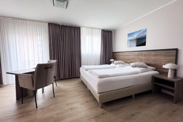 Ein großzügiges modernes Hotelzimmer mit Doppelbett, auf dem weiße und graue Bettwäsche sowie mehrere Zierkissen angeordnet sind. Der helle Holzfußboden erstreckt sich über den gesamten Raum. Links des Betts stehen zwei braune Sessel an einem niedrigen Tisch. Große Fenster mit graubraunen und transparenten Vorhängen bieten viel Licht. An der Wand prangt eine Nordseelandschaftsfotografie. Die Einrichtung kombiniert Komfort mit modernem Design und schafft eine luxuriöse, ruhige Atmosphäre. Ein großzügiges modernes Hotelzimmer mit Doppelbett, auf dem weiße und graue Bettwäsche sowie mehrere Zierkissen angeordnet sind. Der helle Holzfußboden erstreckt sich über den gesamten Raum. Links des Betts stehen zwei braune Sessel an einem niedrigen Tisch. Große Fenster mit graubraunen und transparenten Vorhängen bieten viel Licht. An der Wand prangt eine Nordseelandschaftsfotografie. Die Einrichtung kombiniert Komfort mit modernem Design und schafft eine luxuriöse, ruhige Atmosphäre.