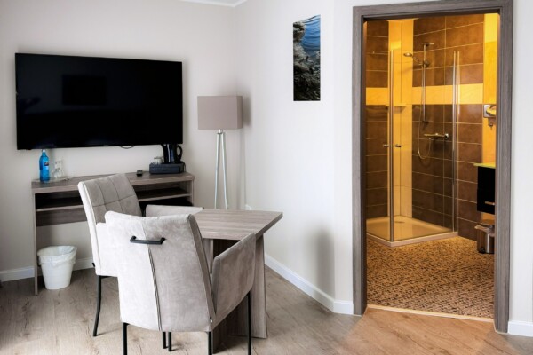 Ein modernes Hotelzimmer mit schwarzem Flachbild-TV an der weißen Wand, darunter ein grauer Schreibtisch mit beigem Stuhl. Rechts öffnet sich eine Badezimmertür zu einer beleuchteten Dusche mit goldenen Fliesen und warmer Beleuchtung. Der hellbraune Holzfußboden zieht sich durch den gesamten Raum. Die offene Badezimmer-Anordnung und moderne Möblierung schaffen ein funktionales, luxuriöses Gesamterlebnis mit zeitgenössischem Design. Ein modernes Hotelzimmer mit schwarzem Flachbild-TV an der weißen Wand, darunter ein grauer Schreibtisch mit beigem Stuhl. Rechts öffnet sich eine Badezimmertür zu einer beleuchteten Dusche mit goldenen Fliesen und warmer Beleuchtung. Der hellbraune Holzfußboden zieht sich durch den gesamten Raum. Die offene Badezimmer-Anordnung und moderne Möblierung schaffen ein funktionales, luxuriöses Gesamterlebnis mit zeitgenössischem Design.
