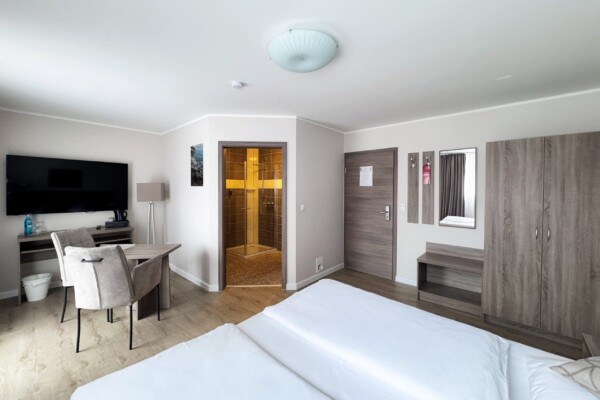 Ein geräumiges modernes Hotelzimmer mit weißem Doppelbett, mehreren Kissen und eleganter Bettwäsche. Breite Fenster mit graubraunen Vorhängen lassen reichlich Licht herein. Der hellbraune Holzfußboden erstreckt sich durchgängig. Links befindet sich ein Arbeitsbereich mit schwarzem TV und grauem Schreibtisch. Eine weiße Deckenleuchte spendet zusätzliches Licht. Die rechte Seite zeigt eine braune Tür und einen Kleiderschrank. Die offene Badezimmertür strahlt goldenes Licht ab. Die Gesamtatmosphäre wirkt komfortabel, funktional und luxuriös. Ein geräumiges modernes Hotelzimmer mit weißem Doppelbett, mehreren Kissen und eleganter Bettwäsche. Breite Fenster mit graubraunen Vorhängen lassen reichlich Licht herein. Der hellbraune Holzfußboden erstreckt sich durchgängig. Links befindet sich ein Arbeitsbereich mit schwarzem TV und grauem Schreibtisch. Eine weiße Deckenleuchte spendet zusätzliches Licht. Die rechte Seite zeigt eine braune Tür und einen Kleiderschrank. Die offene Badezimmertür strahlt goldenes Licht ab. Die Gesamtatmosphäre wirkt komfortabel, funktional und luxuriös.