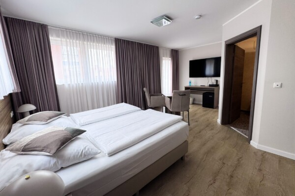 Ein großzügiges Hotelzimmer mit breitem weißem Doppelbett, Kissen in Beige- und Grautönen sowie heller Bettwäsche. Große Fenster mit doppelten Vorhängen in Graubraun und Weiß überfluten den Raum mit Licht. Ein schwarzer Flachbild-TV hängt an der weißen Wand über einem grauen Möbel. Der hellbraune Holzfußboden zieht sich durch alle Bereiche. Links sitzt ein Arbeitsbereich mit braunem Schreibtisch und Stuhl. Die offene Badezimmertür offenbart eine beleuchtete Dusche mit goldenem Glanz. Die minimalistische Farbgebung und hochwertige Ausstattung schaffen eine stressfreie, luxuriöse Luxusatmosphäre. Ein großzügiges Hotelzimmer mit breitem weißem Doppelbett, Kissen in Beige- und Grautönen sowie heller Bettwäsche. Große Fenster mit doppelten Vorhängen in Graubraun und Weiß überfluten den Raum mit Licht. Ein schwarzer Flachbild-TV hängt an der weißen Wand über einem grauen Möbel. Der hellbraune Holzfußboden zieht sich durch alle Bereiche. Links sitzt ein Arbeitsbereich mit braunem Schreibtisch und Stuhl. Die offene Badezimmertür offenbart eine beleuchtete Dusche mit goldenem Glanz. Die minimalistische Farbgebung und hochwertige Ausstattung schaffen eine stressfreie, luxuriöse Luxusatmosphäre.