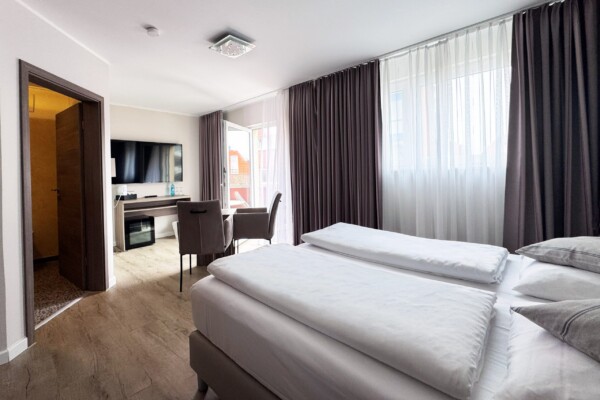 Ein großzügiges modernes Hotelzimmer mit einem breiten Doppelbett, das weiße Bettwäsche und mehrere Zierkissen in Grautönen aufweist. Große Fenster mit graubraunen Vorhängen und weißer Innenschicht fluten den Raum mit Tageslicht. Im Hintergrund links ist eine kleine Kitchenette mit schwarzem Ofen und Kochbereich sichtbar. Ein schwarzer Flachbild-TV hängt an der Wand. Der hellbraune Holzfußboden zieht sich durchgehend. Eine braune Tür öffnet sich zum beleuchteten Badezimmer. Die offene Raumaufteilung und moderne Ausstattung schaffen eine komfortable, funktionale Suite-Atmosphäre. Ein großzügiges modernes Hotelzimmer mit einem breiten Doppelbett, das weiße Bettwäsche und mehrere Zierkissen in Grautönen aufweist. Große Fenster mit graubraunen Vorhängen und weißer Innenschicht fluten den Raum mit Tageslicht. Im Hintergrund links ist eine kleine Kitchenette mit schwarzem Ofen und Kochbereich sichtbar. Ein schwarzer Flachbild-TV hängt an der Wand. Der hellbraune Holzfußboden zieht sich durchgehend. Eine braune Tür öffnet sich zum beleuchteten Badezimmer. Die offene Raumaufteilung und moderne Ausstattung schaffen eine komfortable, funktionale Suite-Atmosphäre.