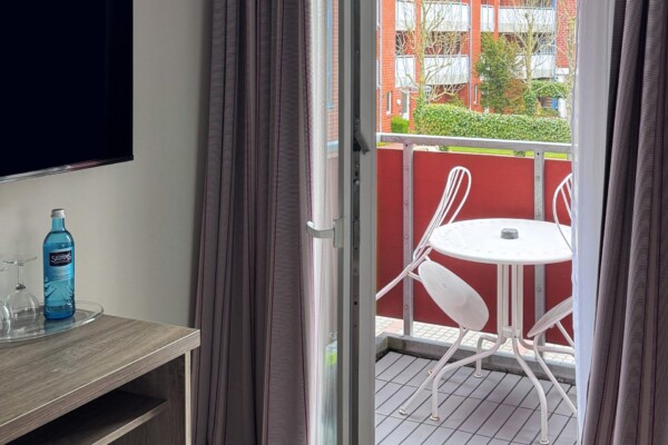 Ein modernes Hotelzimmer mit Zugang zu einem Balkon oder einer Terrasse mit rotem Geländer. Eine große Glasschiebetür bietet freien Blick auf historische rote Backsteinfassaden und grüne Pflanzen außen. Ein schwarzer Flachbild-TV hängt links an der weißen Wand. Ein hölzerner Schreibtisch und moderne Möblierung komplettieren den funktionalen Wohnbereich. Graubraune und weiße Vorhänge rahmen die Aussicht ein. Der hellbraune Holzfußboden und helle Wände schaffen eine lichte, offene Atmosphäre mit direkter Verbindung zur Außenwelt. Ein modernes Hotelzimmer mit Zugang zu einem Balkon oder einer Terrasse mit rotem Geländer. Eine große Glasschiebetür bietet freien Blick auf historische rote Backsteinfassaden und grüne Pflanzen außen. Ein schwarzer Flachbild-TV hängt links an der weißen Wand. Ein hölzerner Schreibtisch und moderne Möblierung komplettieren den funktionalen Wohnbereich. Graubraune und weiße Vorhänge rahmen die Aussicht ein. Der hellbraune Holzfußboden und helle Wände schaffen eine lichte, offene Atmosphäre mit direkter Verbindung zur Außenwelt.