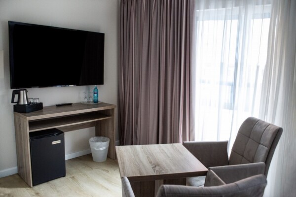 "Ein eleganter Wohnbereich mit großem schwarzem Flachbild-TV an der weißen Wand, darunter ein hölzerner Schreibtisch mit schwarzem Minibar-Kühlschrank und Wasserkaraffe. Im Vordergrund stehen zwei graue Polsterstühle an einem hellholzernen Tisch. Große Fenster mit graubraunen und weißen Doppelvorhängen spenden reichlich Licht. Der hellbraune Laminat-Fußboden erstreckt sich über den gesamten Raum. Die moderne, funktionale Einrichtung kombiniert Arbeits- und Entspannungsbereiche zu einer großzügigen, komfortablen Suite. "Ein eleganter Wohnbereich mit großem schwarzem Flachbild-TV an der weißen Wand, darunter ein hölzerner Schreibtisch mit schwarzem Minibar-Kühlschrank und Wasserkaraffe. Im Vordergrund stehen zwei graue Polsterstühle an einem hellholzernen Tisch. Große Fenster mit graubraunen und weißen Doppelvorhängen spenden reichlich Licht. Der hellbraune Laminat-Fußboden erstreckt sich über den gesamten Raum. Die moderne, funktionale Einrichtung kombiniert Arbeits- und Entspannungsbereiche zu einer großzügigen, komfortablen Suite.