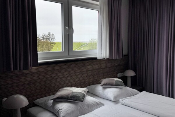 Ein ruhiges modernes Hotelzimmer mit breitem Doppelbett, das weiße Bettwäsche und mehrere graue Zierkissen aufweist. Große Fenster mit graubraunen und weißen Doppelvorhängen bieten einen herrlichen Blick auf grüne Wiesen und ländliche Landschaft. Eine dunkle horizontale Wandpanelierung unterhalb der Fensterbank schafft visuelle Tiefe. Schwarze Nachttische mit weißen Pilzlampen rahmen das Bett ein. Das moderne, minimalistische Design und der Naturausblick schaffen eine friedliche, erholsame Atmosphäre. Ein ruhiges modernes Hotelzimmer mit breitem Doppelbett, das weiße Bettwäsche und mehrere graue Zierkissen aufweist. Große Fenster mit graubraunen und weißen Doppelvorhängen bieten einen herrlichen Blick auf grüne Wiesen und ländliche Landschaft. Eine dunkle horizontale Wandpanelierung unterhalb der Fensterbank schafft visuelle Tiefe. Schwarze Nachttische mit weißen Pilzlampen rahmen das Bett ein. Das moderne, minimalistische Design und der Naturausblick schaffen eine friedliche, erholsame Atmosphäre.