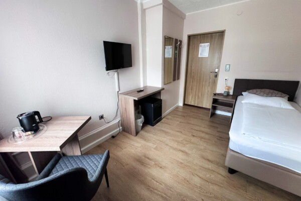 Ein kleines, gepflegtes Hotelzimmer mit einem Einzelbett, einem Schreibtisch und einem Stuhl, einem an der Wand montierten Fernseher, einem Minikühlschrank, einer Holztür, Spiegeln und einem Wasserkocher mit Gläsern auf einem Beistelltisch. Das Zimmer hat helle Wände und einen Bodenbelag in Holzoptik. Ein kleines, gepflegtes Hotelzimmer mit einem Einzelbett, einem Schreibtisch und einem Stuhl, einem an der Wand montierten Fernseher, einem Minikühlschrank, einer Holztür, Spiegeln und einem Wasserkocher mit Gläsern auf einem Beistelltisch. Das Zimmer hat helle Wände und einen Bodenbelag in Holzoptik.