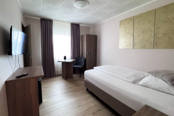 Ein aufgeräumtes, schlichtes Hotelzimmer mit einem Einzelbett, Holzmöbeln, einem Schreibtisch mit Stuhl am Fenster, einem an der Wand montierten Fernseher, einem Kleiderschrank und einem beigen Wandbild über dem Bett. Sonnenlicht fällt durch die hauchdünnen Vorhänge und lässt den Raum hell erscheinen. Ein aufgeräumtes, schlichtes Hotelzimmer mit einem Einzelbett, Holzmöbeln, einem Schreibtisch mit Stuhl am Fenster, einem an der Wand montierten Fernseher, einem Kleiderschrank und einem beigen Wandbild über dem Bett. Sonnenlicht fällt durch die hauchdünnen Vorhänge und lässt den Raum hell erscheinen.