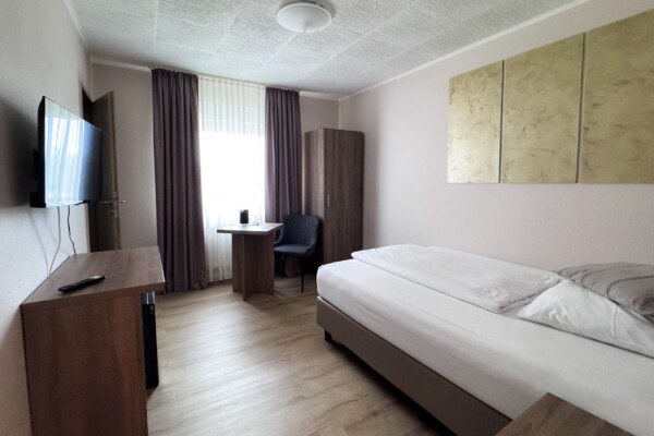 Ein aufgeräumtes, schlichtes Hotelzimmer mit einem Einzelbett, Holzmöbeln, einem Schreibtisch mit Stuhl am Fenster, einem an der Wand montierten Fernseher, einem Kleiderschrank und einem beigen Wandbild über dem Bett. Sonnenlicht fällt durch die hauchdünnen Vorhänge und lässt den Raum hell erscheinen. Ein aufgeräumtes, schlichtes Hotelzimmer mit einem Einzelbett, Holzmöbeln, einem Schreibtisch mit Stuhl am Fenster, einem an der Wand montierten Fernseher, einem Kleiderschrank und einem beigen Wandbild über dem Bett. Sonnenlicht fällt durch die hauchdünnen Vorhänge und lässt den Raum hell erscheinen.