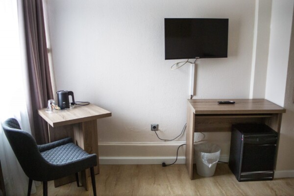 Eine kleine Ecke in einem Hotelzimmer mit zwei Holzschreibtischen, einem gepolsterten Stuhl, einem an der Wand montierten Fernseher, einem Wasserkocher mit Tassen, einem Minikühlschrank, einem Mülleimer, einer Fernbedienung und Stromkabeln; Licht fällt durch ein Fenster auf der linken Seite herein. Eine kleine Ecke in einem Hotelzimmer mit zwei Holzschreibtischen, einem gepolsterten Stuhl, einem an der Wand montierten Fernseher, einem Wasserkocher mit Tassen, einem Minikühlschrank, einem Mülleimer, einer Fernbedienung und Stromkabeln; Licht fällt durch ein Fenster auf der linken Seite herein.
