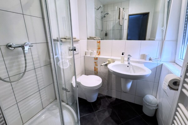 Ein modernes, kompaktes Badezimmer mit weiß gefliesten Wänden, einer Duschkabine aus Glas, einem wandhängenden WC, einem weißen Waschbecken, einem Spiegel über dem Waschbecken, kleinen Ablagen, einem Toilettenpapierhalter, einem Mülleimer und einem Fenster, durch das Tageslicht hereinfällt. Ein modernes, kompaktes Badezimmer mit weiß gefliesten Wänden, einer Duschkabine aus Glas, einem wandhängenden WC, einem weißen Waschbecken, einem Spiegel über dem Waschbecken, kleinen Ablagen, einem Toilettenpapierhalter, einem Mülleimer und einem Fenster, durch das Tageslicht hereinfällt.