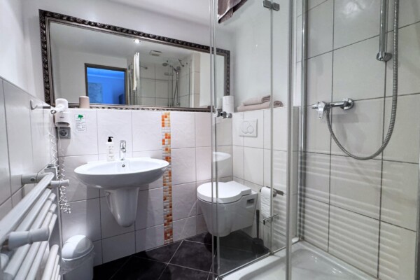 Modernes Badezimmer mit weiß gefliesten Wänden, einer Duschkabine aus Glas, einem wandmontierten Waschbecken und einer Toilette. Über dem Waschbecken befindet sich ein großer Spiegel, links davon ein Handtuchheizkörper, darunter ein kleiner Mülleimer und auf einem Regal über der Toilette gefaltete Handtücher. Modernes Badezimmer mit weiß gefliesten Wänden, einer Duschkabine aus Glas, einem wandmontierten Waschbecken und einer Toilette. Über dem Waschbecken befindet sich ein großer Spiegel, links davon ein Handtuchheizkörper, darunter ein kleiner Mülleimer und auf einem Regal über der Toilette gefaltete Handtücher.