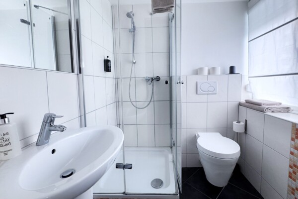 Das moderne, weiß geflieste Badezimmer verfügt über eine verglaste Duschkabine, ein wandhängendes WC, ein weißes Waschbecken mit einer verchromten Armatur, Handtücher, einen Seifenspender, Toilettenpapierrollen sowie Tageslicht, das durch ein Fenster mit einer weißen, durchsichtigen Jalousie hereinfällt. Das moderne, weiß geflieste Badezimmer verfügt über eine verglaste Duschkabine, ein wandhängendes WC, ein weißes Waschbecken mit einer verchromten Armatur, Handtücher, einen Seifenspender, Toilettenpapierrollen sowie Tageslicht, das durch ein Fenster mit einer weißen, durchsichtigen Jalousie hereinfällt.