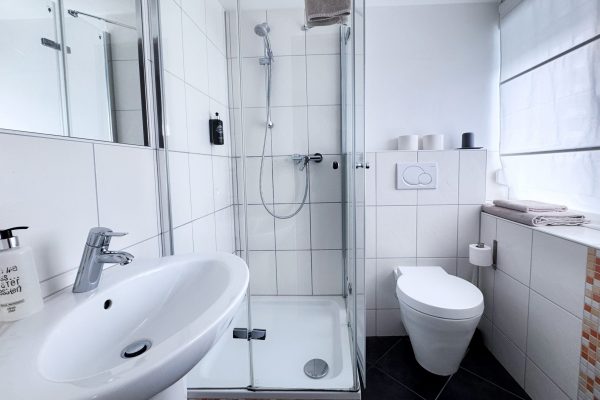Das moderne, weiß geflieste Badezimmer verfügt über eine verglaste Duschkabine, ein wandhängendes WC, ein weißes Waschbecken mit einer verchromten Armatur, Handtücher, einen Seifenspender, Toilettenpapierrollen sowie Tageslicht, das durch ein Fenster mit einer weißen, durchsichtigen Jalousie hereinfällt. Das moderne, weiß geflieste Badezimmer verfügt über eine verglaste Duschkabine, ein wandhängendes WC, ein weißes Waschbecken mit einer verchromten Armatur, Handtücher, einen Seifenspender, Toilettenpapierrollen sowie Tageslicht, das durch ein Fenster mit einer weißen, durchsichtigen Jalousie hereinfällt.