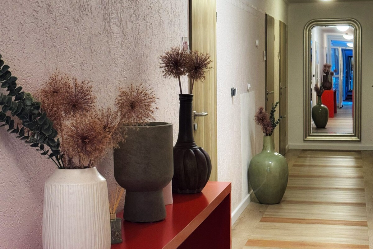 Ein eleganter moderner Hotelflur mit strukturierter altrosa-farbener Wandverkleidung. Eine rote moderne Bank oder Podium präsentiert stilvolle schwarze und weiße Deko-Vasen, gefüllt mit getrockneten Gräsern und Pampasgrass. Eine dunkelgrüne Keramik-Vase setzt einen natürlichen Akzent. Der hellbraune Holzfußboden erstreckt sich durchgehend. Mehrere cremefarbene Hotelzimmertüren säumen die rechte Seite. Ein großer Wandspiegel reflektiert das warme Licht. Die durchdachte Stilisierung verbindet moderne Ästhetik mit gemütlicher Hotellichkeit.