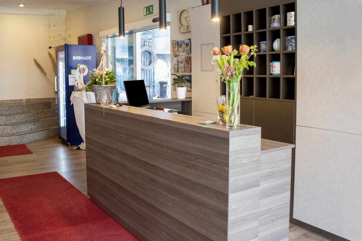 Ein einladender moderner Hotel-Empfangsbereich mit großzügigem graubraunem Holz-Empfangsthesen. Ein roter Eingangsteppich kontrastiert mit dem hellen Laminat-Fußboden. Frische Schnittblumen in transparenten Glas-Vasen — mit rosa Tulpen und grüner Floristik — begrüßen Gäste. Moderne schwarze Pendelleuchten aus Metall erhellen den Raum. Ein Fenster mit Außenblick, die Bionade-Getränkestation und ein modernes Bücherregal mit Dekoelementen komplettieren die Rezeption. Ein Laptop auf dem Tresen signalisiert professionelle Hotelstandards. Die warme, moderne Gestaltung schafft eine herzliche Ankunftserfahrung.