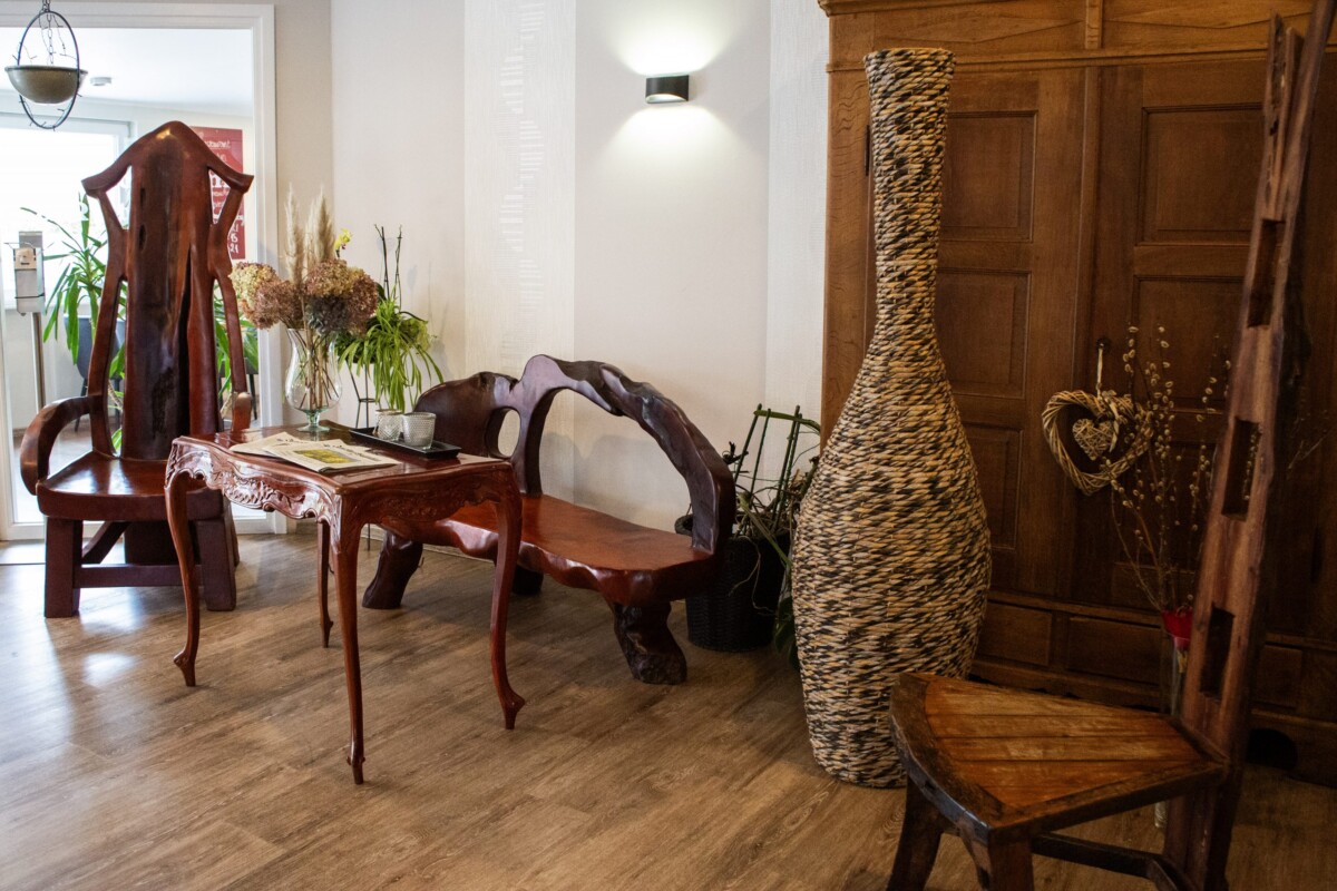 Eine elegante Hotellobby mit klassischen Holzmöbeln in warmer Rotbraun-Tönung. Ein großes Fenster mit Ausblick und hängender Metalldekor-Korb fügt Leichtigkeit hinzu. Mehrere Stühle mit geschwungener Rückenlehne laden zum Verweilen ein. Frische Grünpflanzen und getrocknete Blüten in Glasvasen bringen Natur ins Interieur. Eine große spiralförmige Weidenkorb-Säule dient als dekoratives Element. Rechts öffnen sich dunkelbraune Holztüren. Der hellbraune Holzfußboden und warm weiße Wandflächen schaffen eine klassisch-elegante, entspannende Atmosphäre mit zeitlosem Charme.