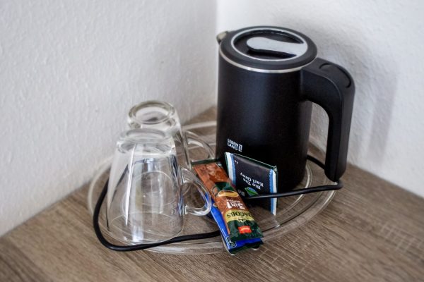 Auf einem Glastablett stehen ein schwarzer Wasserkocher, zwei umgedrehte Glastassen, eine Packung Instantkaffee und zwei Teebeutel. Das Tablett steht auf einer Holzfläche in einer Zimmerecke in der Nähe weißer Wände. Auf einem Glastablett stehen ein schwarzer Wasserkocher, zwei umgedrehte Glastassen, eine Packung Instantkaffee und zwei Teebeutel. Das Tablett steht auf einer Holzfläche in einer Zimmerecke in der Nähe weißer Wände.