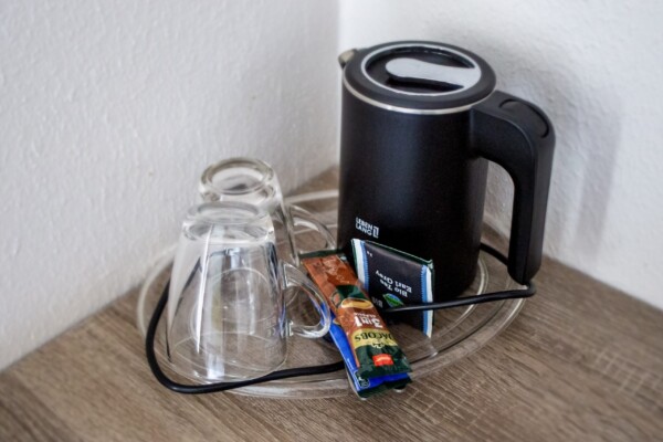 Auf einem Glastablett stehen ein schwarzer Wasserkocher, zwei umgedrehte Glastassen, eine Packung Instantkaffee und zwei Teebeutel. Das Tablett steht auf einer Holzfläche in einer Zimmerecke in der Nähe weißer Wände. Auf einem Glastablett stehen ein schwarzer Wasserkocher, zwei umgedrehte Glastassen, eine Packung Instantkaffee und zwei Teebeutel. Das Tablett steht auf einer Holzfläche in einer Zimmerecke in der Nähe weißer Wände.