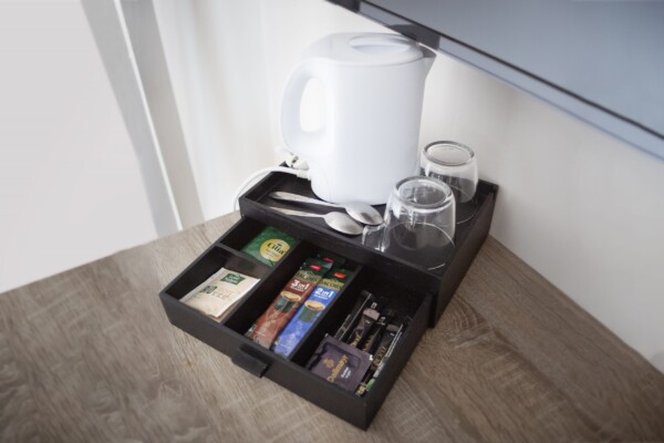 Eine Getränkestation in einem Hotelzimmer mit einem weißen Wasserkocher, zwei umgedrehten Gläsern, zwei Löffeln, Teebeuteln, Instantkaffee-Sticks, Milchpulver, Zucker und einer kleinen Praline, alles ordentlich auf einem schwarzen Tablett auf einer Holzunterlage angeordnet. Eine Getränkestation in einem Hotelzimmer mit einem weißen Wasserkocher, zwei umgedrehten Gläsern, zwei Löffeln, Teebeuteln, Instantkaffee-Sticks, Milchpulver, Zucker und einer kleinen Praline, alles ordentlich auf einem schwarzen Tablett auf einer Holzunterlage angeordnet.