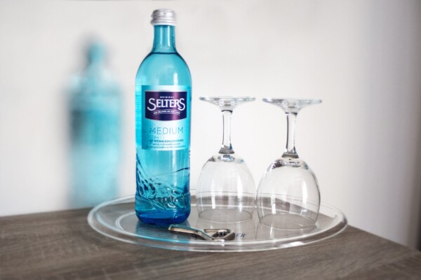 Auf einem durchsichtigen, runden Tablett steht eine blaue Glasflasche mit Selters-Mineralwasser neben zwei umgedrehten Weingläsern und einem Flaschenöffner aus Metall; das Ganze befindet sich auf einer Holzunterlage, im Hintergrund ist eine verschwommene Flasche zu sehen. Auf einem durchsichtigen, runden Tablett steht eine blaue Glasflasche mit Selters-Mineralwasser neben zwei umgedrehten Weingläsern und einem Flaschenöffner aus Metall; das Ganze befindet sich auf einer Holzunterlage, im Hintergrund ist eine verschwommene Flasche zu sehen.