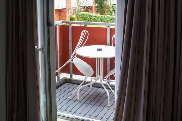 Durch eine offene Glastür mit flatternden Vorhängen blickt man auf einen kleinen Balkon. Auf dem Balkon steht ein runder weißer Metalltisch mit zwei passenden Stühlen, von denen einer hervorgezogen ist. Das Geländer ist einheitlich rot, und draußen sind Wohnhäuser und Grünflächen zu sehen. Durch eine offene Glastür mit flatternden Vorhängen blickt man auf einen kleinen Balkon. Auf dem Balkon steht ein runder weißer Metalltisch mit zwei passenden Stühlen, von denen einer hervorgezogen ist. Das Geländer ist einheitlich rot, und draußen sind Wohnhäuser und Grünflächen zu sehen.