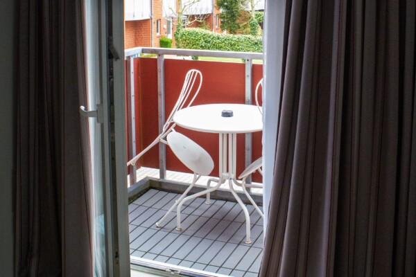 Durch eine offene Glastür mit flatternden Vorhängen blickt man auf einen kleinen Balkon. Auf dem Balkon steht ein runder weißer Metalltisch mit zwei passenden Stühlen, von denen einer hervorgezogen ist. Das Geländer ist einheitlich rot, und draußen sind Wohnhäuser und Grünflächen zu sehen. Durch eine offene Glastür mit flatternden Vorhängen blickt man auf einen kleinen Balkon. Auf dem Balkon steht ein runder weißer Metalltisch mit zwei passenden Stühlen, von denen einer hervorgezogen ist. Das Geländer ist einheitlich rot, und draußen sind Wohnhäuser und Grünflächen zu sehen.