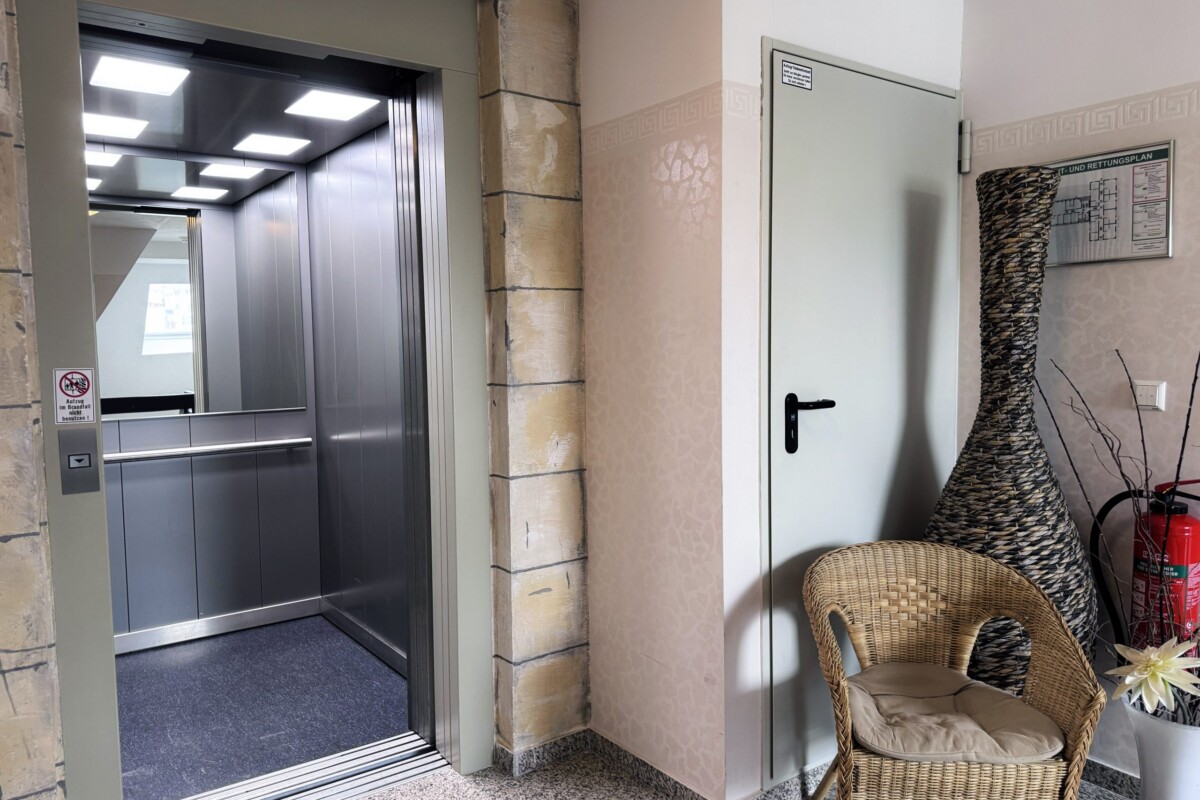 Ein moderner Hotelflur mit glänzendem Aufzug, dessen spiegelnde graue Edelstahl-Fassade das Licht reflektiert. Die Wand ist in warmer Beige-Creme-Tönung gestrichen. Eine große dekorative Weidenkorb-Säule mit Flechtwerk-Details prägt das Ambiente. Ein beiger Weidenkorb-Stuhl mit beigem Kissen bietet eine gemütliche Sitzgelegenheit. Eine schlichte weiße Tür rechts und graubrauner Fliesenboden komplettieren den funktionalen Raum. Die dezente, stilvolle Dekoration und moderne Aufzugtechnik schaffen einen einladenden, gehobenen Hotelstandard.