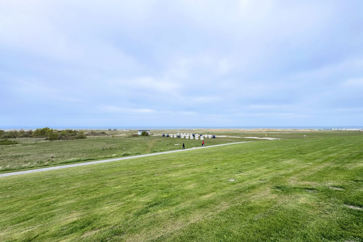 Eine malerische flache Nordseelandschaft mit einer grünen Wiese, die sich endlos zum Horizont erstreckt. Ein gepflasterter grauer Weg führt geradeaus in die Ferne und verbindet die Strandanlage. Am Horizont sind mehrere farbige Strandkörbe in einer Reihe sichtbar. Zwei Fußgänger spazieren gemächlich auf dem Weg. Der klare, helle Himmel mit einzelnen weißen Wolken spiegelt sich in der friedlichen Atmosphäre. Die weitläufige, flache Topografie und grüne Vegetation verkörpern die entspannte Urlaubscharme der Nordseeklimazone.