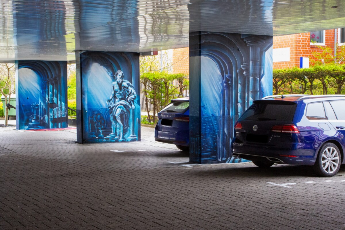 Ein künstlerisch gestalteter Parkplatz-Unterstand mit mehreren dunkelblaugetönten Säulen, die mit klassischen mythologischen Wandmalereien bemalt sind. Die Kunstwerke zeigen Neptun-ähnliche Figuren und architektonische Details in Hellblau und Weiß. Mehrere moderne Autos in Blau und Schwarz parken geschützt unter der Konstruktion. Das rote Backsteingebäude des Hotels und grüne Vegetation bilden den Hintergrund. Die thematisch konsistente, künstlerisch hochwertige Gestaltung verbindet Funktionalität mit kulturellem Anspruch und schafft ein unvergessliches Parkplatz-Erlebnis.