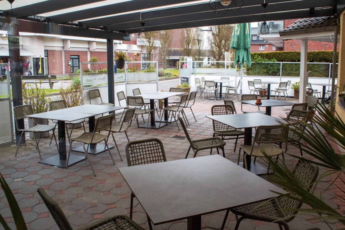 Eine großzügige moderne Restaurantterrasse unter elegantem schwarzem Metallrahmen-Dach mit transparenten Lichtpanelen. Mehrere Esstische sind mit zeitgenössischen graubraunen Korbstühlen bestuhlt. Die roten und braunen Pflastersteine bilden einen warmen Untergrund. Große Fensterfront mit Sichtschutzplatte grenzt die Terrasse zur Straße ab. Rote Backsteingebäude und grüne Hecken prägen das urbane Umfeld. Eine grüne Gartenanlage mit Vegetation belebt die Szenerie. Der offene, moderne Terrassenstil verbindet Komfort mit zeitgenössischem Design und lädt zu angenehmen Mahlzeiten ein.