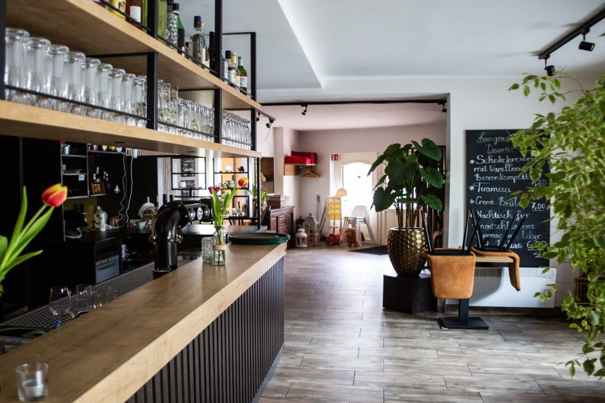 Eine professionelle modern gestaltete Bar-Lounge mit intelligenter Raumnutzung: Ein langer heller Holztresen mit schwarzem Gitterwerk-Panel bildet die Basis. Darüber erstrecken sich großflächige Holzregale mit professioneller Bar-Ausstattung — transparente Cocktail-Gläser, Likörflaschen und Barausstattung sind elegant arrangiert. Eine schwarze strukturierte Rückwand schafft Kontrast. Grüne Topfpflanzen und eine handgeschriebene Speisekarte auf schwarzer Tafel verleihen natürliche, gemütliche Akzente. Die moderne, funktionale Gestaltung verbindet professionelle Standards mit einladender Atmosphäre.