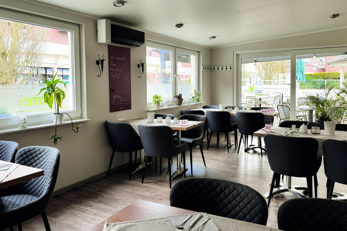 Ein hell gestalteter Restaurantbereich mit großflächigen Fenstern, die direkten Blick auf die Außenterrasse und Landschaft ermöglichen. Schwarze moderne Polsterstühle mit Quilting-Details sitzen an Holztischen mit Nummernkennzeichnung. Eine dunkle Tafel an der Wand zeigt Speisen und Preise. Grüne Topfpflanzen bringen Natur ins Innere. Die cremefarbenen Wände und schwarzen Akzente schaffen einen modernen, gehobenen Standard. Die großzügige Fensterfront und offene Gestaltung vermitteln Transparenz und Helligkeit.