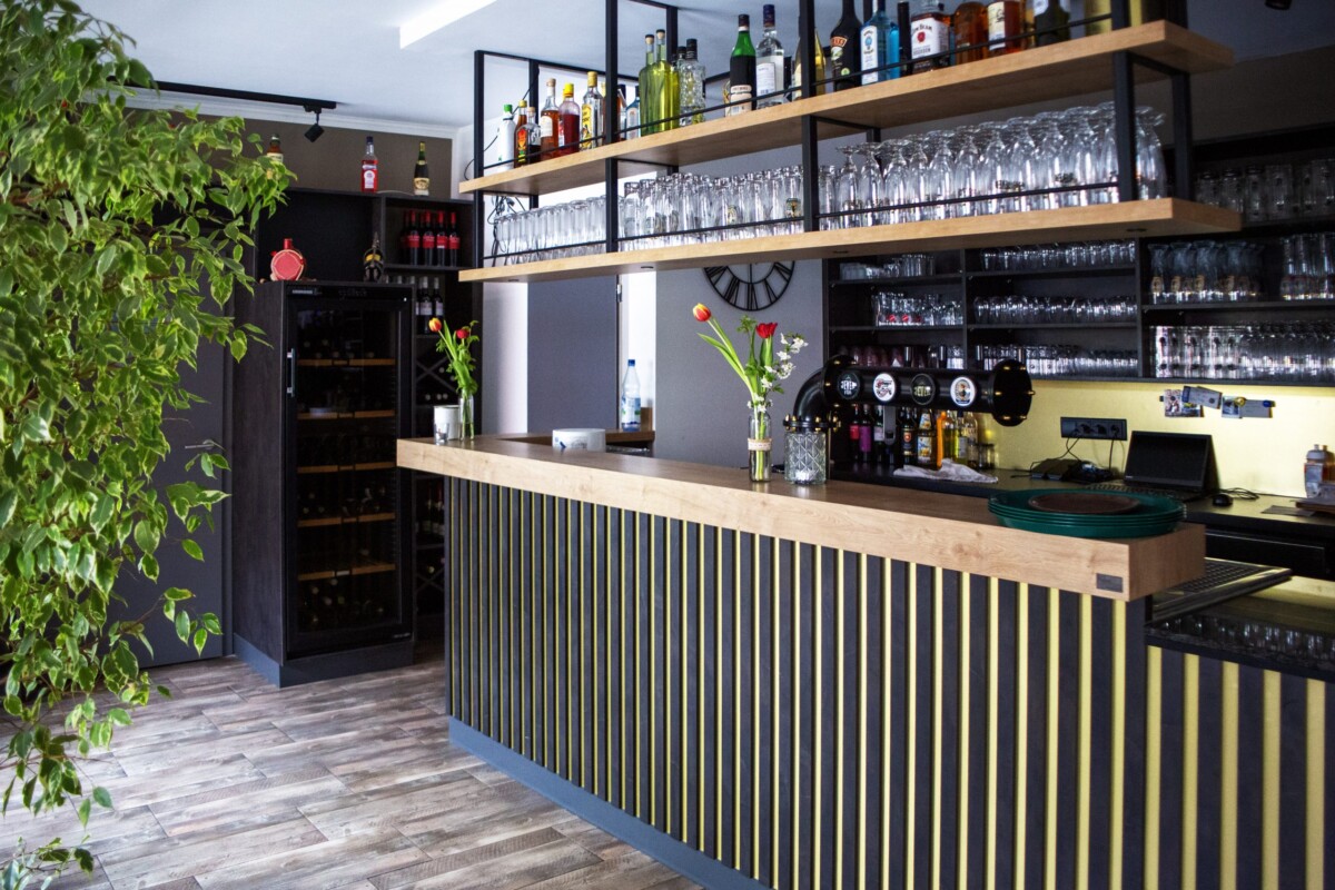 Eine schick gestaltete moderne Bar mit breitem hellholzernem Tresen, dessen Front mit modernem schwarzem Gitterwerk-Panel gestaltet ist. Große hölzerne Wandregale zeigen eine exklusive Auswahl an Spirituosen und Likören. Transparent Gläser, Besteck und Barausstattung sind übersichtlich arrangiert. Eine lebendige grüne Topfpflanze links belebt den Bereich. Die dunkle Wandfläche dahinter schafft Kontrast. Moderne Beleuchtung und hochwertige Bar-Möblierung vermitteln professionelle Gastlichkeit und gehobenes Ambiente.