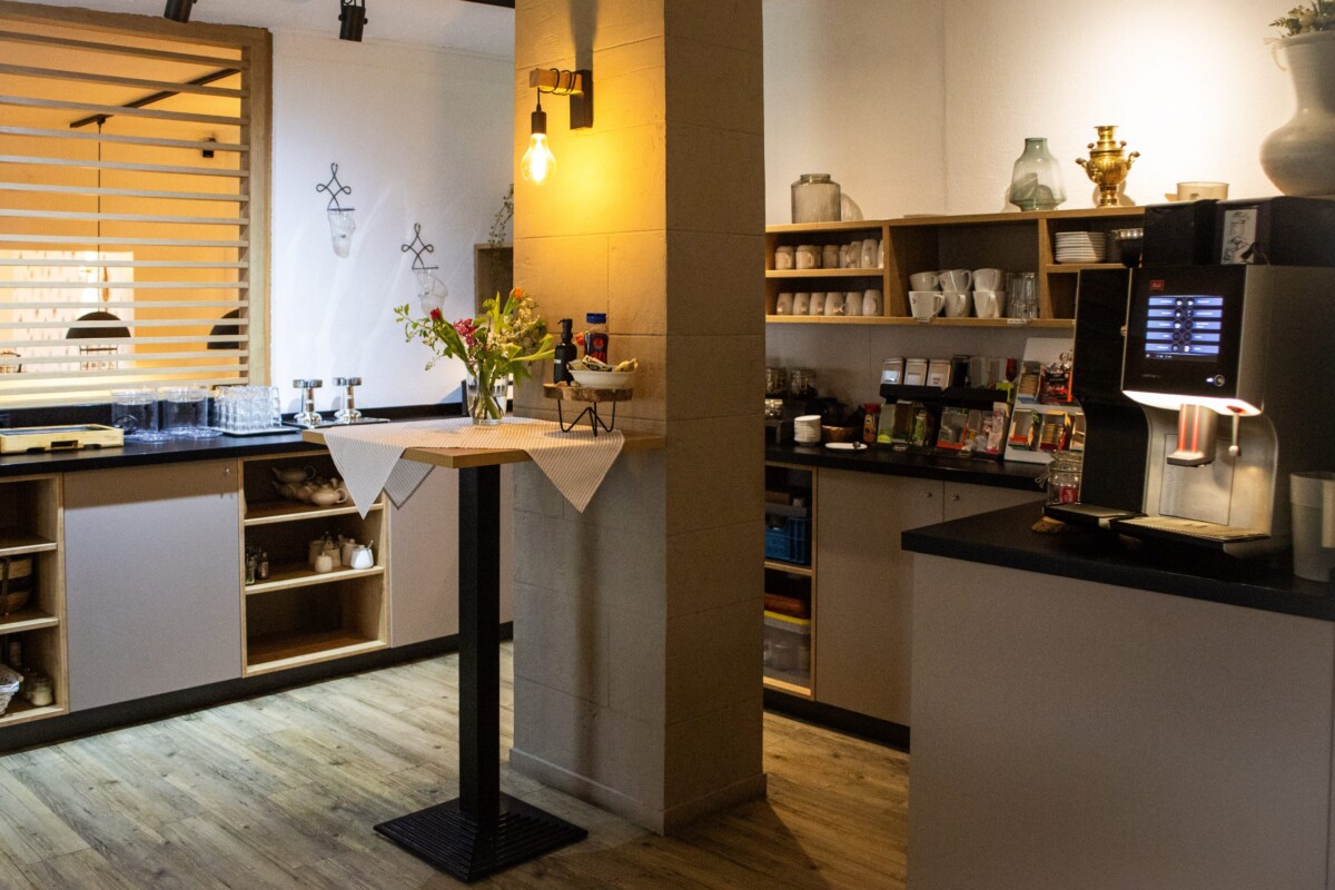Ein stilvoller Café-Lounge-Bereich mit warmem Holztresen und großflächigen schwarzen Wandregalen. Elegante goldfarbene Metallgestelle und dekorative Objekte (Vasen, Flaschen) prägen das Ambiente. Eine farbenfrohe Topfpflanze mit roten und gelben Blüten bringt Leben ins Design. Fenster mit orangen/gelben Lamellen-Rollos schaffen warme Lichtstimmung. Ein schwarzer Säulenelement und Holzfußboden vervollständigen die moderne Ästhetik. Die warme Farbgebung und durchdachte Dekoration schaffen eine einladende, gehobene Café-Atmosphäre.